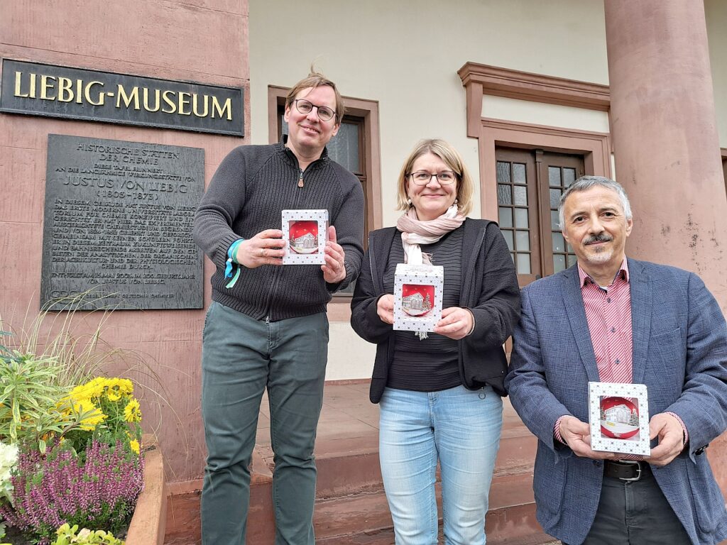 Die Gießener Weihnachtskugel mit dem Liebig-Museum als Motiv wird von drei Personen präsentiert.